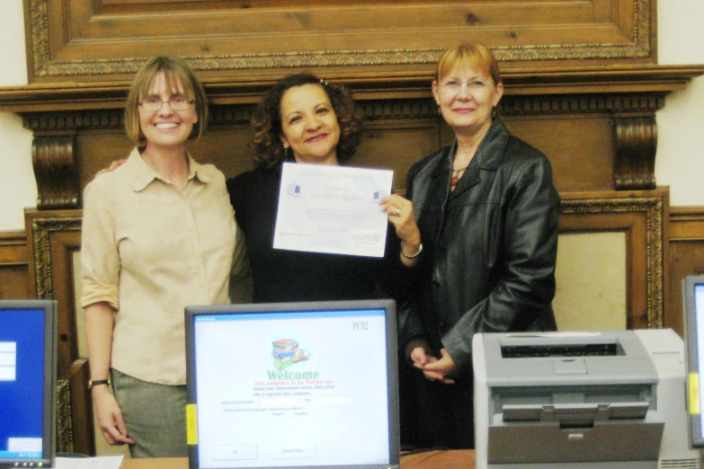 three women smile while holding a paper next to their computers