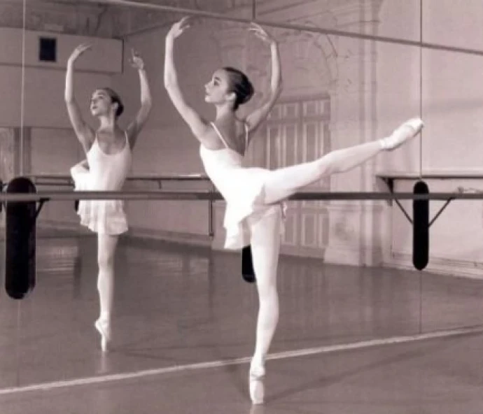 two ballerinas standing and one is stretching with their arms up