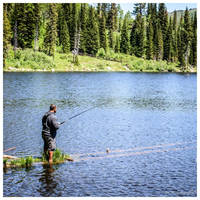 a man fishing in the water with trees and mountains behind