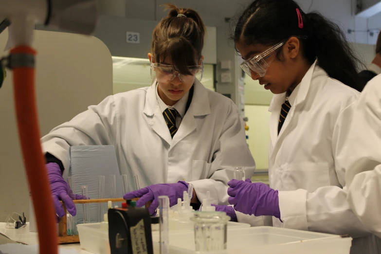 two girls dressed in lab coats stand in front of pipe test tubes, beaks and beakers