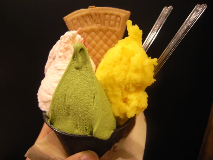 a hand holding a cup filled with three different colored ice creams