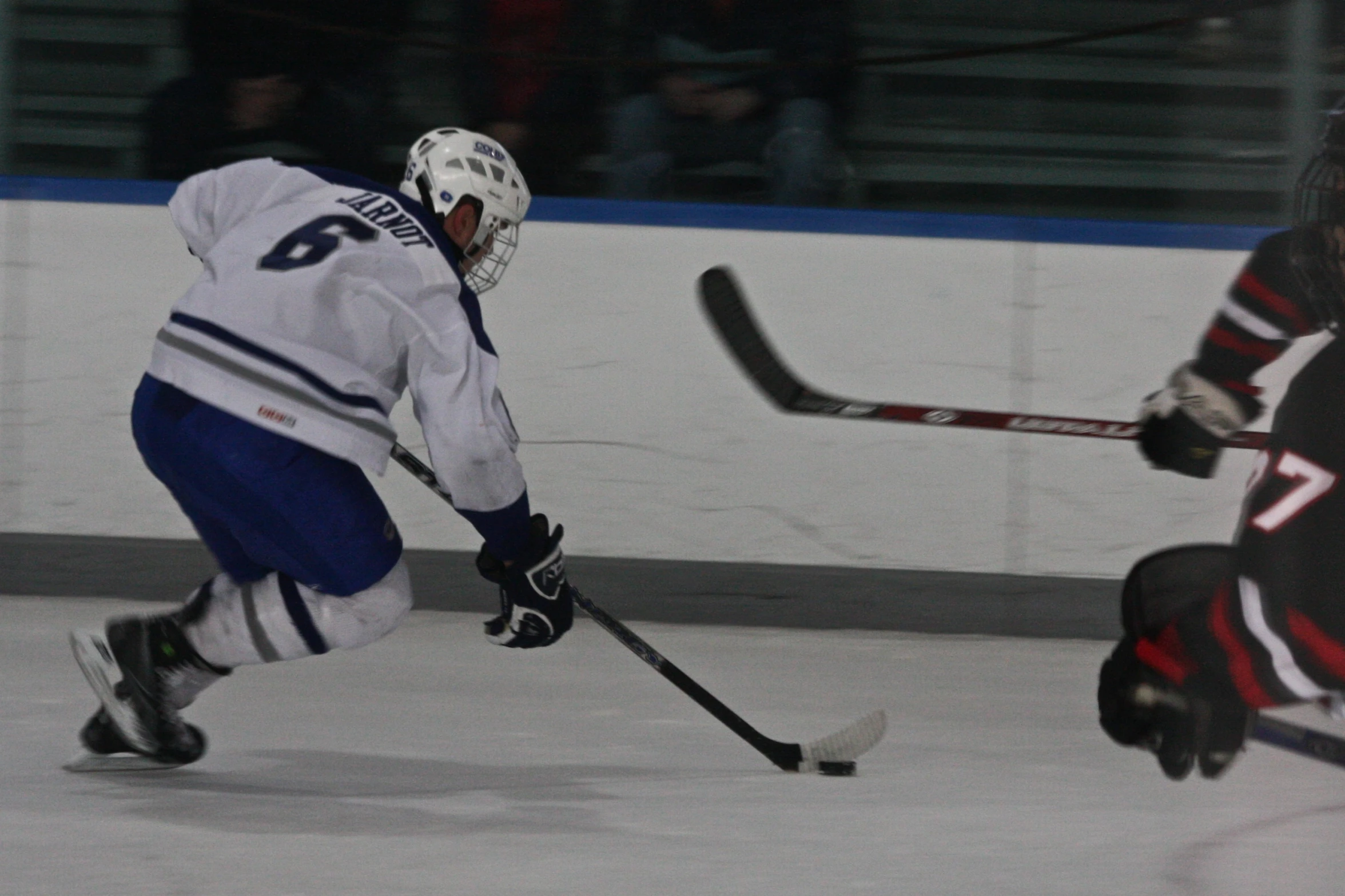 an ice hockey player with a white jersey and blue and white jersey moves toward the puck as another player wears black and red hockey boots