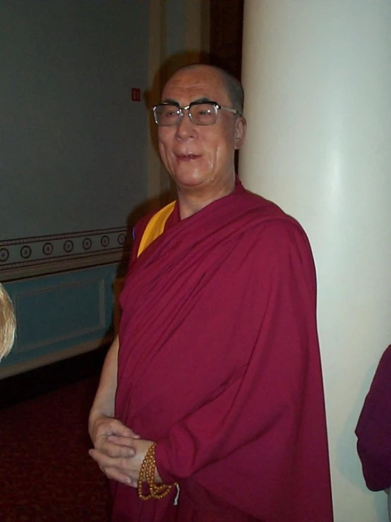 an older man in a red monk's outfit, stands next to a pillar