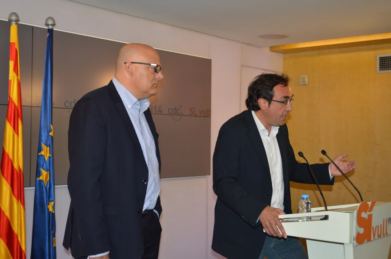 two men speaking at a podium with flags behind them