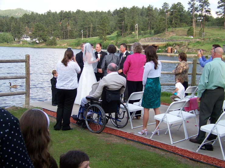 a wedding ceremony at a lake with a man in a wheelchair and the bride and groom walking along the walkway