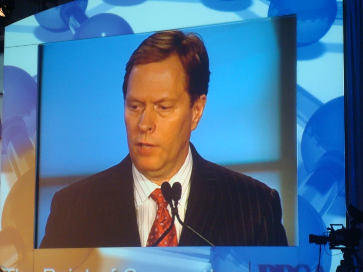 the man is speaking to a crowd in front of the large screen