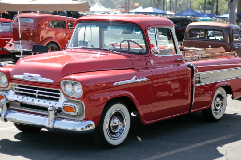 an older, red, pickup truck sits parked near some other antique trucks