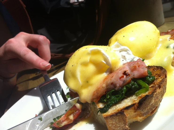 a person holds the knife near a sandwich with eggs, bacon and spinach