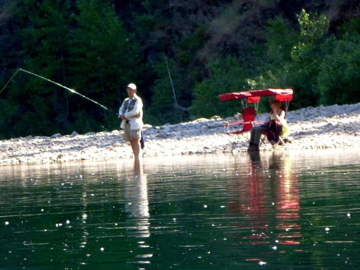 a man with a fishing pole stands in the water as another person stands on the side holding onto a fishing pole