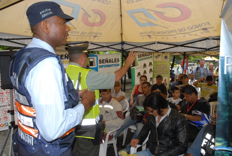 a person in a hat and vest stands under a tent while talking to a group
