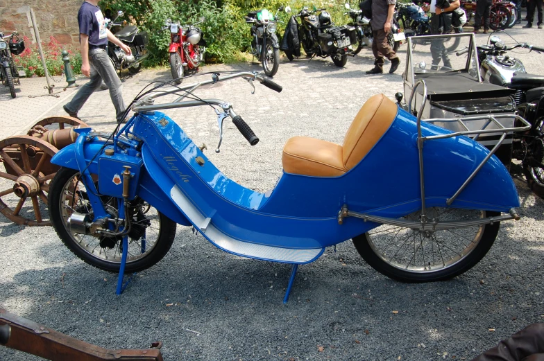 a blue motor bike sits in gravel with people in the background