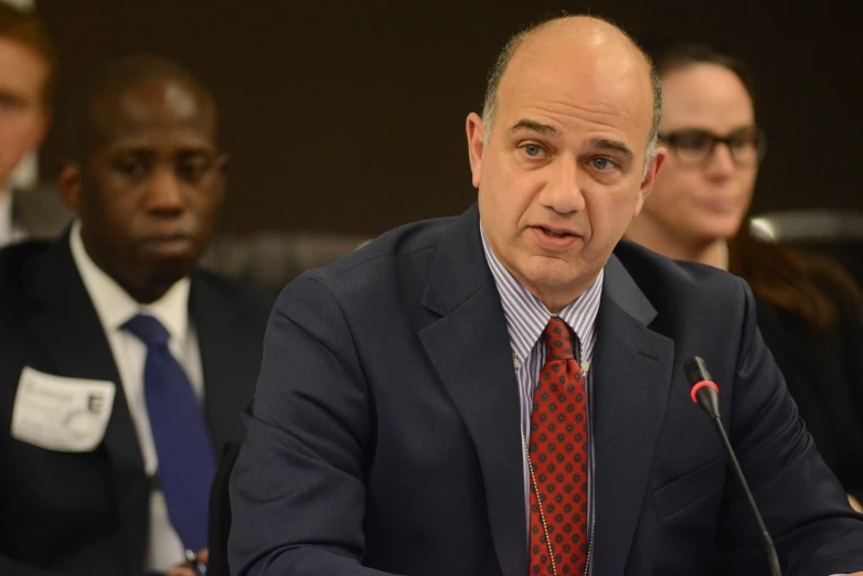 a man speaking while sitting at a table with other people behind him