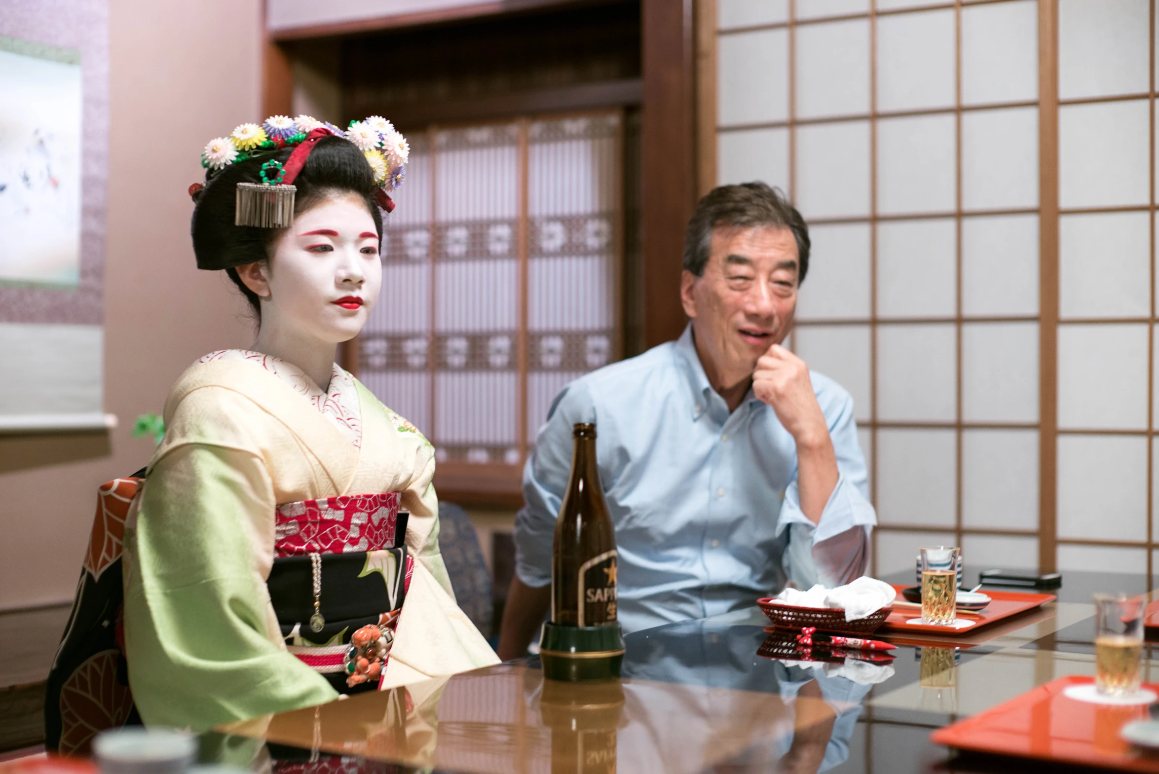 a man sitting at a table with a geisha woman looking on