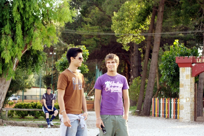 two boys are standing in a park, one with a skateboard