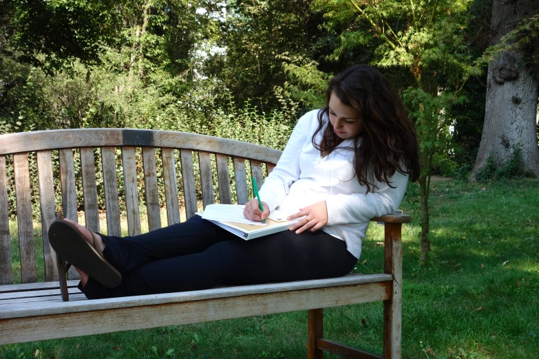 a young lady laying on a park bench writes in a notebook