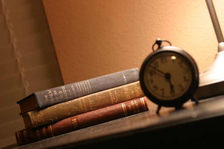 an alarm clock next to books with a lamp behind it