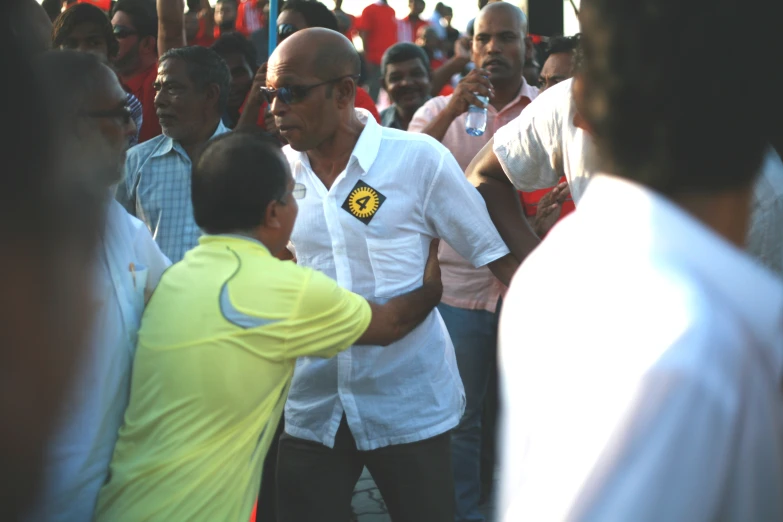 a man with a white shirt and green t - shirt standing near a crowd of people