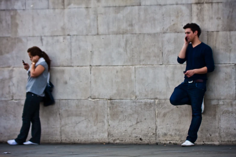 there is a man and woman standing near a wall talking on the cell phones