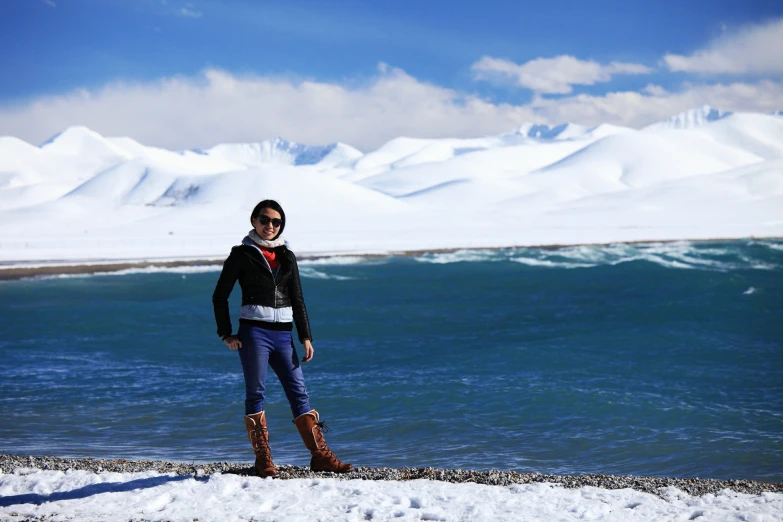 a person standing on the edge of a body of water with a large amount of snow on the mountains