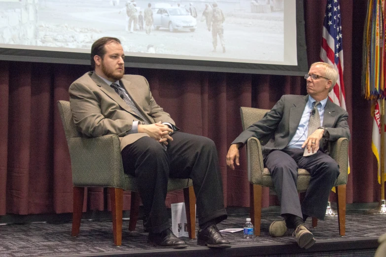 two men in suits sit in chairs and look at the screen