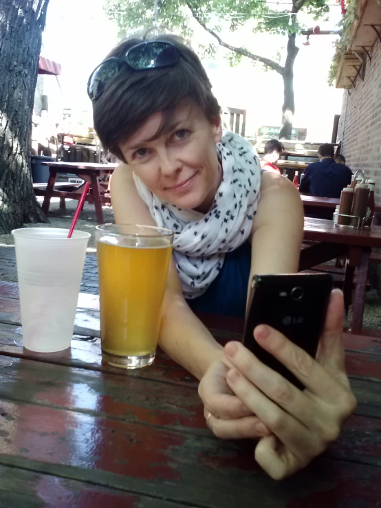 a girl sits at a table holding a cell phone, next to a beer