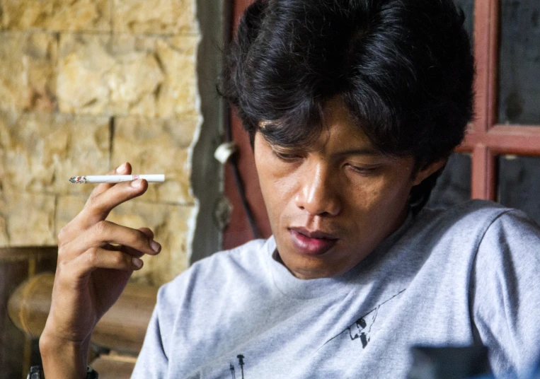 an asian man holding a cigarette looking down while smoking