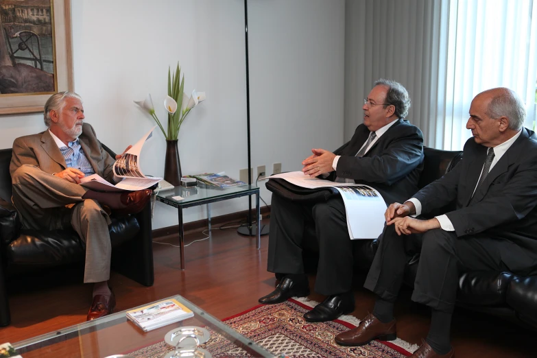 two men and one is sitting in chairs while holding papers