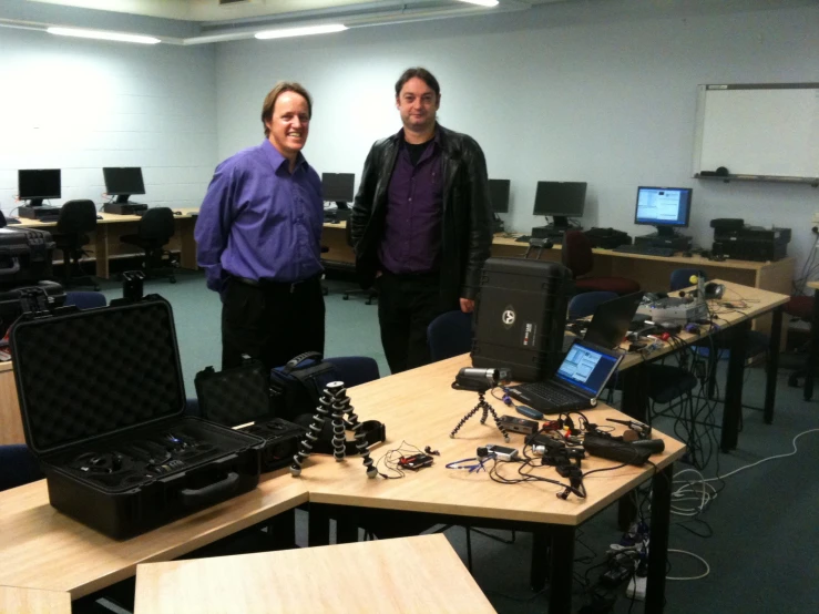 two men are standing in a room that has desks with laptops on it