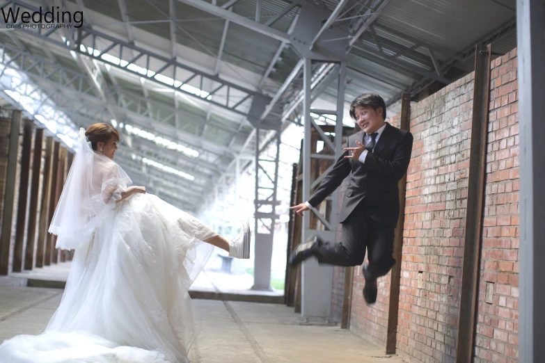 a woman in a wedding dress leaps from a red brick wall to a man in a tuxedo
