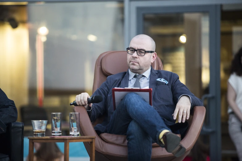 a man in glasses and a suit sits in a chair with an ipad