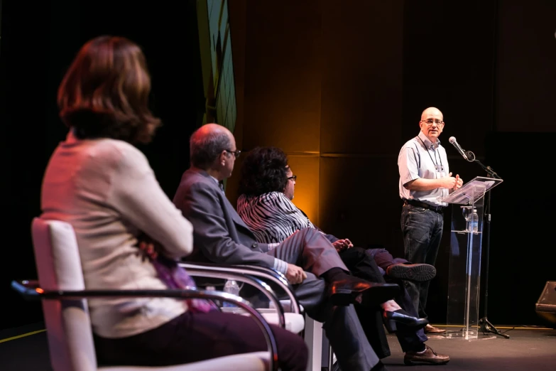 people seated on chairs with a man giving a speech