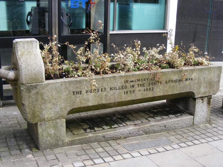 a gray cement bench on the street has flowers in a planter