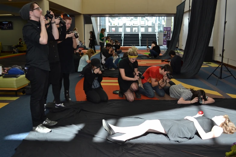 a group of people sit and stand around a sleeping man in a studio