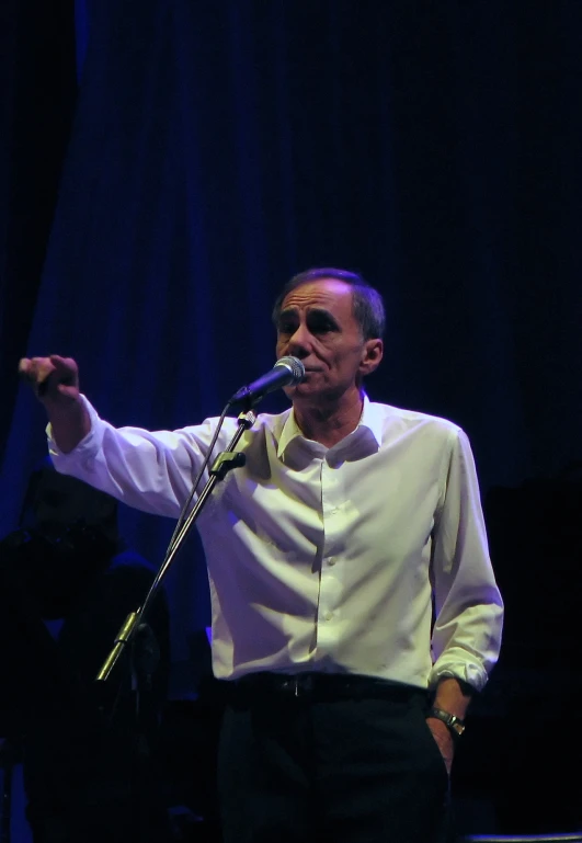 man in white shirt and black pants holding microphone with hands out while standing on stage