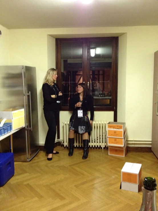 two women sit in a small room near a refrigerator