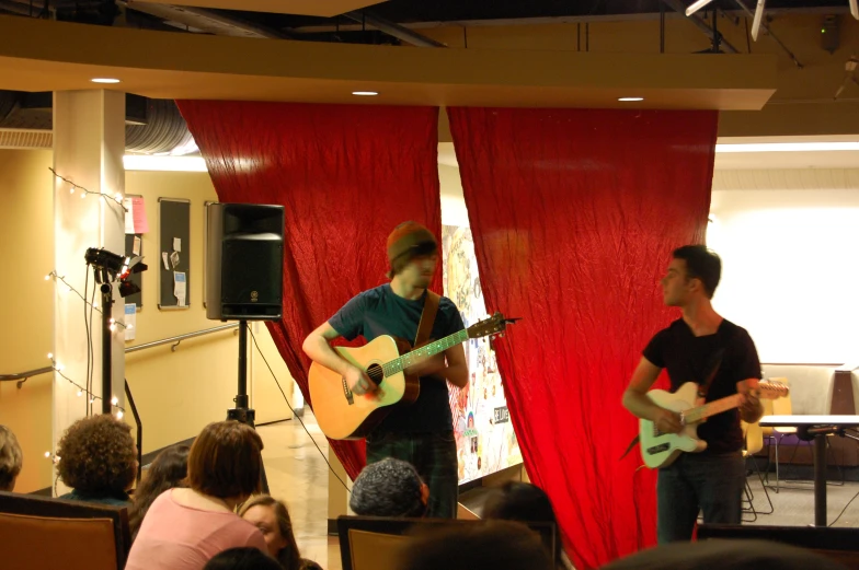two young men are playing acoustic music in a room with people seated