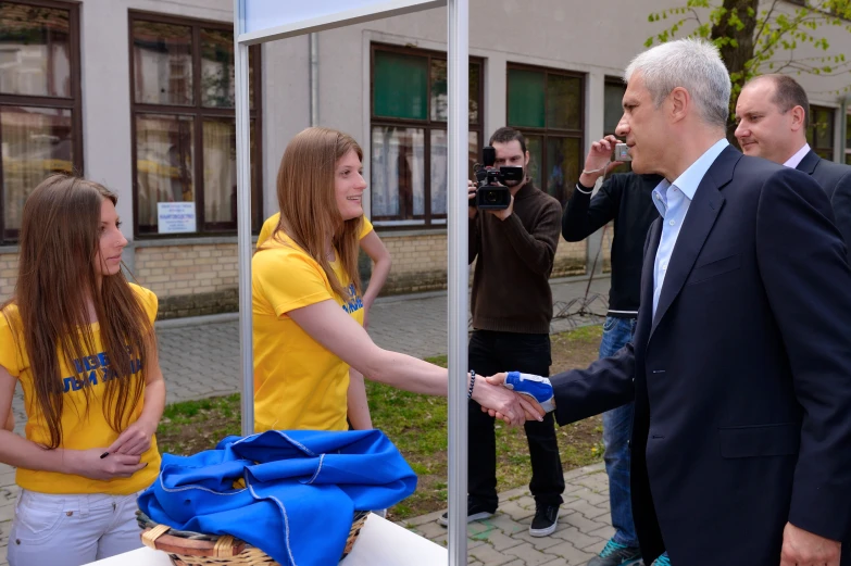 two women shake hands near a man in a suit