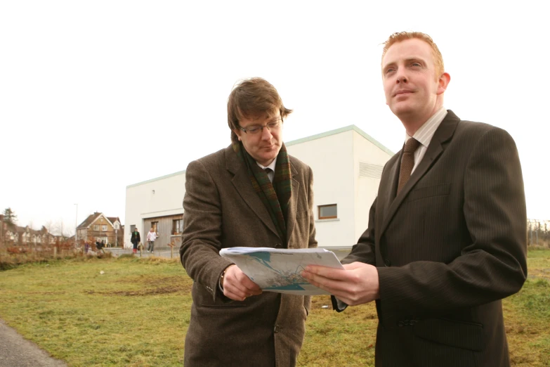 two men are standing in a yard looking at a piece of paper