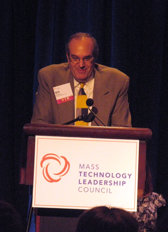 man at podium smiling in front of microphone with banner on side