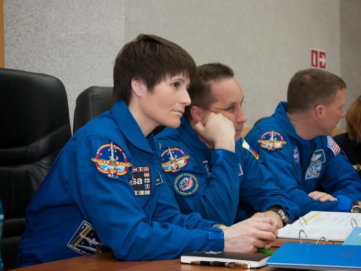 three people sitting at a desk in blue spacesuits