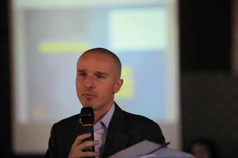 man in business suit talking into microphone while standing in front of screen