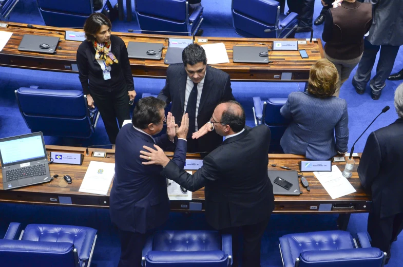 a group of men in suits standing around a room filled with blue chairs