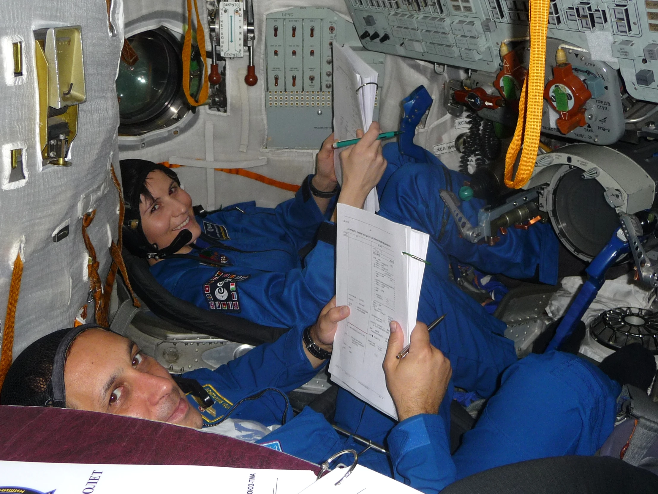 two astronauts in the capsule looking through a piece of paper