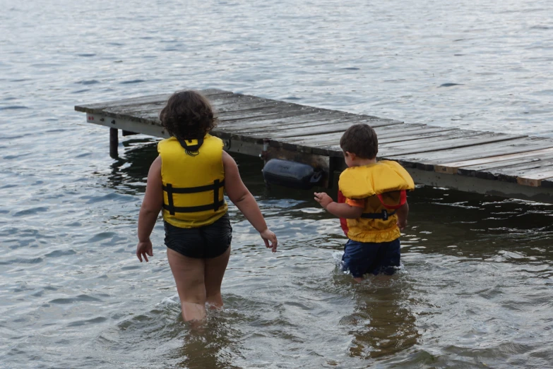 two boys wearing life jackets walk into the water towards a dock