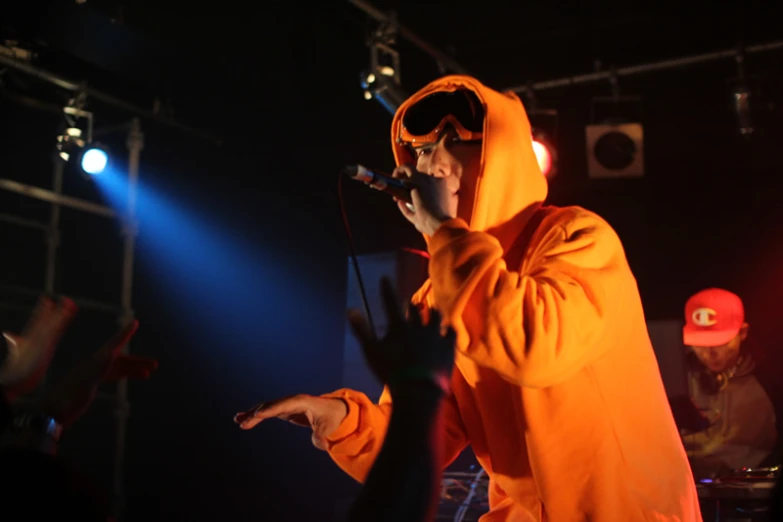 a young man wearing an orange hooded suit sings into his microphone while surrounded by other people