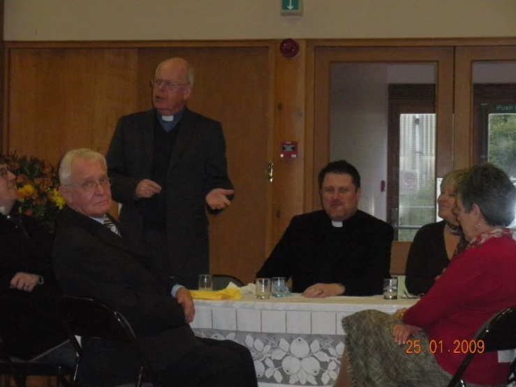 a priest standing at a table while several people sit at the tables