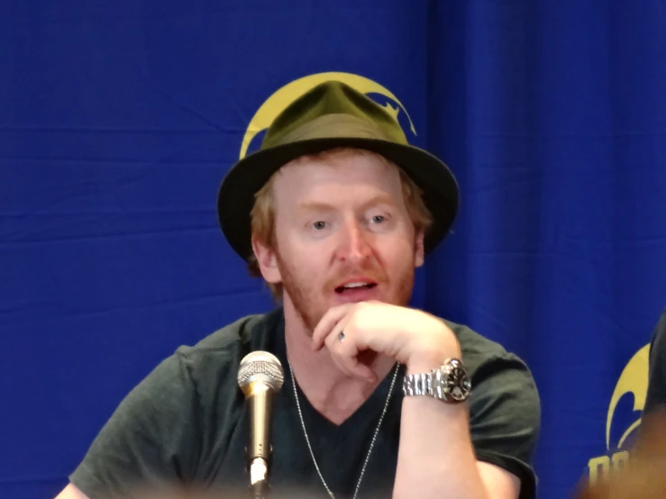a man wearing a green shirt, hat and silver rings is sitting at a table with a microphone