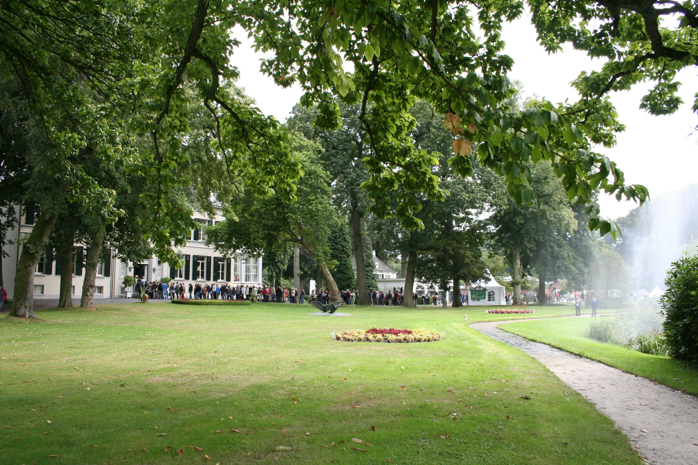 a grassy area has trees, a path and water shooting from a fountain