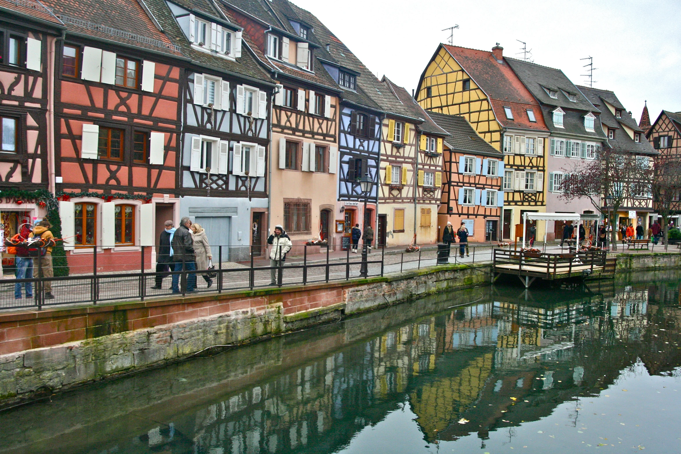 people walking on the boardwalk next to a river with buildings along the side