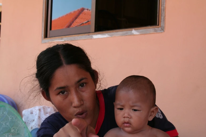 woman holding baby pointing at camera while looking outside